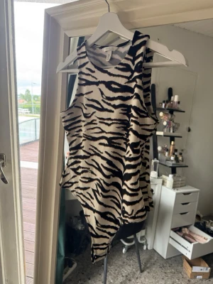 Zebramönstrad ärmlös body HM - Säljer en snygg zebramönstrad body i svart, brun och vit. Bodyn är ärmlös med hög hals och har en asymmetrisk, croppad passform som ger en edgy look. Perfekt för dig som vill sticka ut med ett djurmönster och trendig siluett.