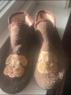 Bruna tofflor med hundmotiv(barn) - Ett par bruna tofflor i mjukt tyg med broderade hundansikten framtill. Tofflorna har en högre kant runt ankeln och svart sula. Insidan är beige och kanten har en mörkare brun nyans. Perfekta för att hålla fötterna varma hemma.