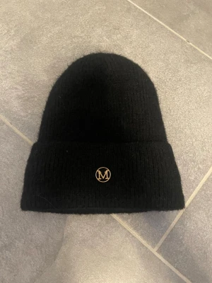 Svart ribbstickad mössa med M-logga - Säljer en svart ribbstickad mössa med uppvikt kant och en liten guldfärgad M-logga framtill. Klassisk och enkel design som passar till det mesta. Materialet ser ut att vara mjukt och värmande, perfekt för kyliga dagar. Passar för barn mellan 2-5 år. 
