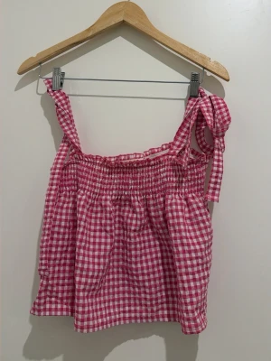 Rutig rosa linnetopp från DF Young - Söt rosa och vit rutig linnetopp från DF Young i storlek XS. Toppen har smock upptill, axelband med knyt och ett luftigt, avslappnat fall. Perfekt för varma dagar och ger en fräsch sommarkänsla.