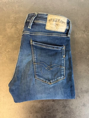 Blå Replay Anbass jeans, stl 32/34 - Säljer ett par klassiska blå jeans från Replay med smal passform och snygga slitningar. Jeansen har fem fickor, ljusare tvättade partier och Replay-logga på bakfickan. Perfekta till vardagslooken och tillverkade i mjukt denimtyg.