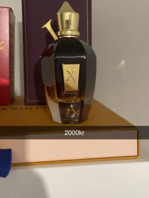 Lyxig parfymflaska med gulddetaljer - Exklusiv parfym i mörk glasflaska med guldfärgat lock och dekorativ X på framsidan. Flaskan har en elegant, kantig form och känns riktigt lyxig. Perfekt för dig som vill ha något unikt och snyggt på parfymhyllan.
