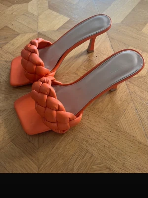 Orange flätade pumps med klack - Snygga orange pumps med fyrkantig tå och flätad rem över foten. Skorna har öppen häl och en smal klack som ger en elegant look. Tillverkade i ett mjukt material som ser ut som konstläder. Perfekta för dig som vill sticka ut.
