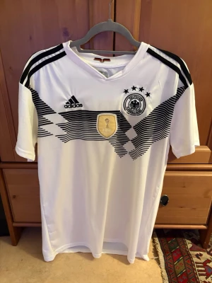 Tysklands matchtröja Adidas herr - Vit Tyskland matchtröja från Adidas med svarta detaljer och tre stjärnor ovanför landslagets emblem. Tröjan har svart mönster på bröstet, tre svarta ränder på axlarna och ett guldigt VM-märke i mitten. Tillverkad i lätt och ventilerande material.