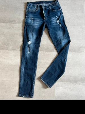 Mörkblå slitna jeans med raka ben - Säljer ett par mörkblå jeans med raka ben och slitningar framtill för en cool, avslappnad look. Jeansen har klassisk femficksdesign och knappgylf. Materialet är denim i bomull. Perfekta för dig som gillar en trendig streetstil.