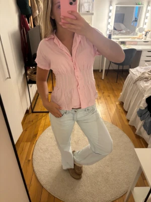 Ljusrosa kortärmad blus med knappar - Söt ljusrosa blus med korta ärmar och knappar framtill. Blusen har en figurnära passform med rynkade detaljer längs midjan och en klassisk krage. Perfekt att styla med jeans för en avslappnad look.