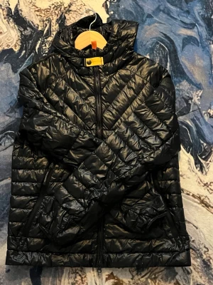 Svart dunjacka med huva - Snygg svart dunjacka med huva och dragkedja framtill. Jackan har ett quiltat mönster i chevron-stil och två sidofickor med dragkedja. Perfekt för kalla vinterdagar och ger en sportig look.