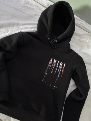 Svart hoodie - Amiri  - Säljer en asfet Svart Amiri hoodie med stor huva och snörning. På bröstet och ryggen finns ett vitt AMIRI-tryck med färgstänk i rött och blått som rinner neråt. Hoodien har en klassisk känguruficka och är tillverkad i mjukt material.