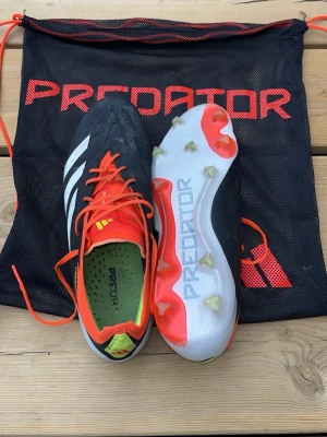 Adidas Predator fotbollsskor svart/orange - Säljer ett par Adidas Predator fotbollsskor i svart med vita ränder och orange detaljer. Skorna har snörning, grön innersula och fasta dobbar för spel på naturgräs. Snygg design med Predator-logga på sulan och medföljande svart förvaringspåse.