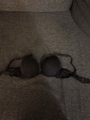 Svart push-up bh med spetsdetaljer - Säljer en svart push-up bh med vadderade kupor och fina spetsdetaljer på framsidan. Axelbanden är justerbara och bh:n har hakknäppning bak. Materialet är en mix av spets och slät textil för en snygg look.
