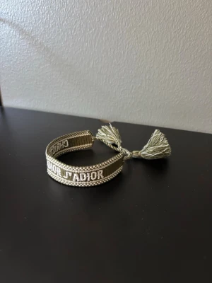 Grön broderat armband Dior - Snyggt beige broderat armband från Dior med texten 'Christian Dior J'Adior' i vitt. Armbandet har justerbara tofsar i ändarna och är gjort i textilmaterial. Perfekt accessoar för att lyfta din outfit.
