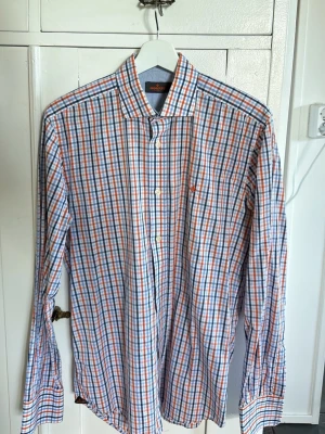 Rutig långärmad skjorta i bomull - Säljer en Morrus rutig skjorta i vitt, blått och orange. Skjortan har lång ärm, knappar framtill och en liten broderad detalj på bröstet. Tillverkad i mjuk bomull och passar perfekt till jeans eller chinos. 