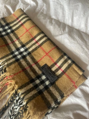 Beige rutig halsduk från Burberry - Klassisk beige halsduk från Burberry i mjuk ull med det ikoniska rutmönstret i svart, vitt och rött. Halsduken har fransar i ändarna och känns både tidlös och trendig. Perfekt accessoar för att lyfta vilken outfit som helst.