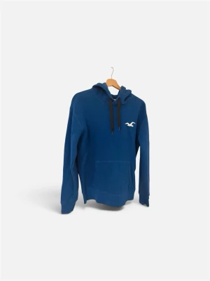 Blå hoodie från Hollister - Blå hoodie från Hollister med vit logga på bröstet och stort tryck med märkesnamnet längs ena ärmen. Klassisk känguruficka framtill och svarta dragsnören i huvan. Perfekt för en avslappnad och cool stil.