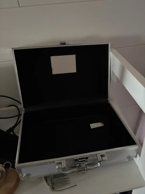Snygg sminkväska i aluminium - Praktisk och stilren sminkväska i silverfärgat aluminium med spegel på insidan av locket. Perfekt för att förvara smink, borstar och andra skönhetsverktyg. Väskan har ett robust handtag och metallspänne för säker förslutning.