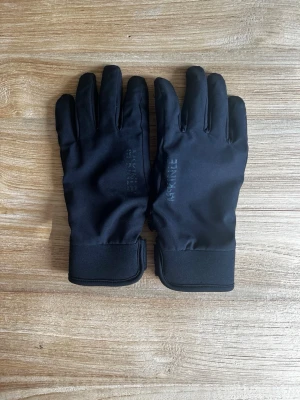 Svarta sportiga handskar med kardborre - Säljer ett par svarta handskar med justerbar kardborrestängning vid handleden och greppvänliga partier i handflatan. Handskarna har en slimmad passform och är tillverkade i ett syntetmaterial som passar perfekt för utomhusaktiviteter under kyligare dagar.