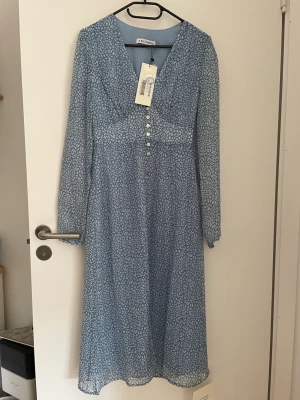 ADOORE paris dress stl 36 - Söt blå långklänning med små vita blommor, v-ringad hals och dekorativa knappar framtill. Klänningen har lång ärm och ett lätt, luftigt tyg som passar perfekt till varmare dagar. Stilren och feminin design.
