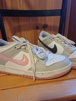 Nike sneakers med rosa och svart  - Snygga Nike sneakers i low-modell med vit bas, ljusgrå detaljer och olika färgade swoosh-loggor – ena skon har rosa och den andra svart. Skorna har klassisk perforerad tå och vita skosnören. Yttermaterialet är en mix av läder och syntet.