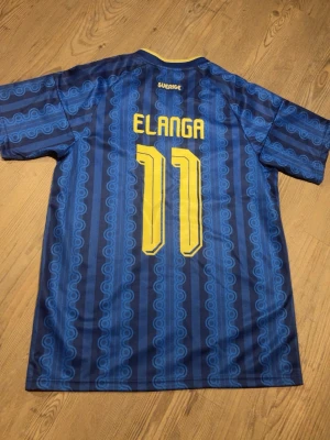 Blå Sverige fotbollströja Elanga 11 - Säljer en blå fotbollströja med gula detaljer från Sverige, Elanga 11 på ryggen. Tröjan har mönstrade ränder, gula axelband och krage samt landslagsemblem på bröstet. Perfekt för dig som vill stötta landslaget med stil. Kom med prisförslag 