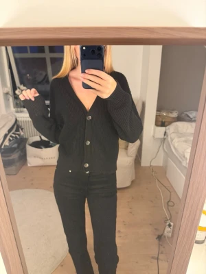 Svart ribbad kofta med knappar - Säljer en svart ribbstickad kofta med v-ringning och stora silvriga knappar framtill. Koftan har lång ärm och en avslappnad passform som passar perfekt till jeans eller kjol. Materialet ser mjukt och mysigt ut, perfekt för kyligare dagar.
