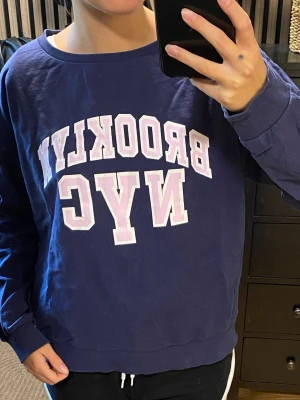 Lilablå sweatshirt med BROOKLYN NYC-tryck - Lilablå tunn sweatshirt med vit och ljusrosa BROOKLYN NYC-text framtill. Tröjan har rund halsringning, långa ärmar och ribbade muddar. Tröjan är lite för stor för mig och därav vill jag sälja den.