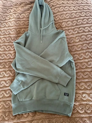 Grön hoodie från Jack & Jones - Snygg grön hoodie från Jack & Jones med klassisk huva och dragsko. Tillverkad i mjukt bomullsmaterial och har en liten svart logotyp-patch på nedre delen. Perfekt för en chill och avslappnad stil. Storlek S/M? Herr storlek
