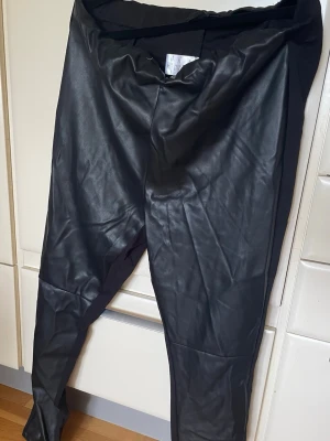 Svarta Mywear leggings i fuskskinn - Snygga svarta Mywear woman leggings i fuskskinn med stretchiga partier på sidorna för extra komfort. De har en hög midja och smal passform som ger en cool och trendig look. Perfekta att matcha med oversized tröjor eller crop tops för en edgy stil.