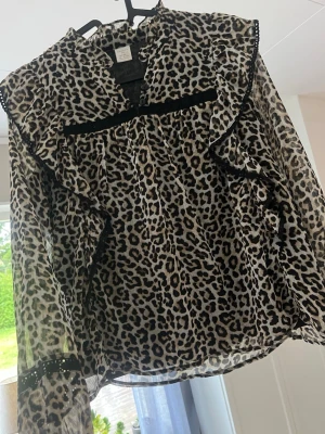 Leopardmönstrad blus med volanger - Snygg blus med leopardmönster och transparenta långärmar. Volangdetaljer framtill och svart band över bröstet ger en trendig look.  Materialet är tunt och lätt, vilket gör blusen luftig och bekväm.