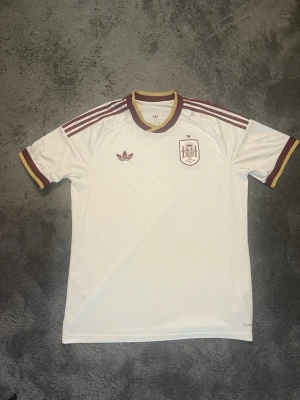 Borta tröja VM Spanien fotbollströja - Säljer en vit Spanien fotbollströja från Adidas med vinröda detaljer. Tröjan har korta ärmar, tre klassiska ränder på axlarna och ett subtilt mönster i tyget. Spanien-emblemet och Adidas-loggan finns på tröjan. Köpt på unisport i Stockholm. Skriv om du har funderingar!