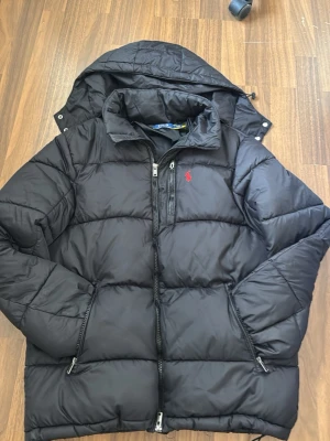 Polo Ralph Lauren - pufferjacka - Snygg svart pufferjacka från Polo Ralph Lauren med röd logga på bröstet. Jackan har dragkedja framtill, bröstficka med dragkedja och två sidofickor. Tillverkad i glansigt material och har en avtagbar huva. Perfekt för kalla vinterdagar. 
