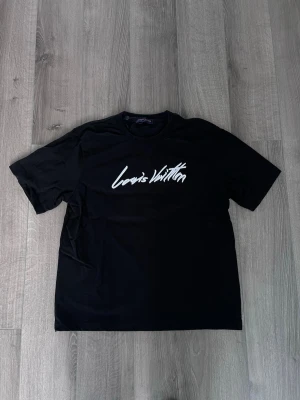 Svart Louis Vuitton t-shirt - Snygg svart t-shirt med Louis Vuitton-logga i vitt framtill. Klassisk passform med korta ärmar och rund hals. Perfekt till jeans eller shorts för en clean och stilren look.