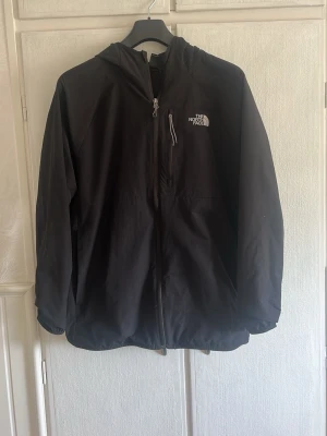 Svart vindjacka från The North Face - Står att det är L men sitter mer som en M Snygg svart vindjacka från The North Face med huva och dragkedja framtill. Jackan har logga på bröstet och ryggen, samt en praktisk bröstficka med dragkedja. Perfekt för blåsiga dagar och enkel att matcha med allt.
