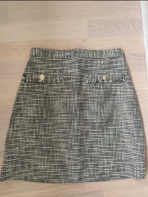 Kort rutig kjol med knappar - Säljer en kort kjol i svart och vitt med rutigt mönster. Kjolen har två dekorativa fickor framtill med guldfärgade knappar och är gjord i ett tjockare material som känns lite ulligt. Perfekt för dig som gillar klassisk och trendig stil. Använt en gång från H&M