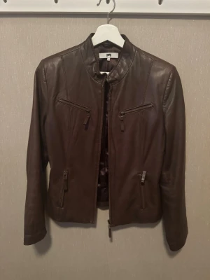 Mörkbrun skinnjacka med dragkedjor - Snygg mörkbrun skinnjacka med ståkrage och fyra dragkedjeförsedda fickor framtill. Jackan har en figurnära passform och stilrena sömdetaljer som ger en modern look. Perfekt för dig som vill ha en klassisk men trendig jacka i garderoben. 100% läder, nypris 1000 kr