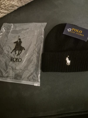 Svart ribbstickad mössa Polo Ralph Lauren - Snygg svart ribbstickad mössa från Polo Ralph Lauren med klassisk vit broderad logga framtill. Mössan har uppvikt kant och är tillverkad i mjukt material som håller dig varm och stilren under kalla dagar.