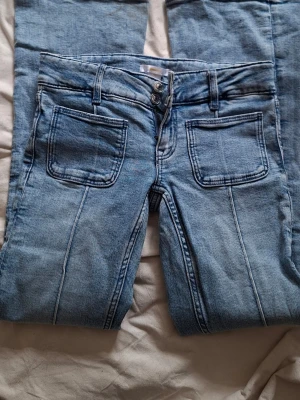 Ljusblå jeans med utsvängda ben - Säljer ett par ljusblå jeans med utsvängda ben och dubbla sömmar framtill. Jeansen har stora fyrkantiga framfickor, bakfickor och tre knappar i gylfen. Klassisk denimlook i mjukt material, perfekta till vardagsstilen.