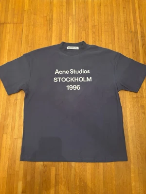 Mörkblå Acne Studios t-shirt - Snygg mörkblå t-shirt från Acne Studios med vit text 'Acne Studios STOCKHOLM 1996' på bröstet. Klassisk passform och rund halsringning. Perfekt för dig som gillar stilrena och trendiga plagg.