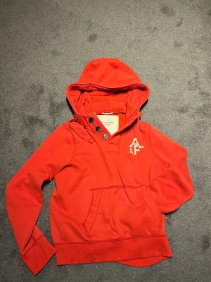 Röd hoodie från Abercrombie & Fitch - Snygg röd hoodie med huva och knappar vid halsen från Abercrombie & Fitch. Stor magficka framtill och logga i vitt på bröstet. Perfekt för en avslappnad och cool stil.