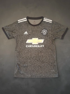Manchester United bortatröja Pogba - Säljer en grå Manchester United bortatröja från Adidas med Pogba och nummer 6 på ryggen. Tröjan har ett vågigt mönster, vita detaljer och klubbmärke på bröstet. Tillverkad i lätt funktionsmaterial, perfekt för fotboll eller träning.