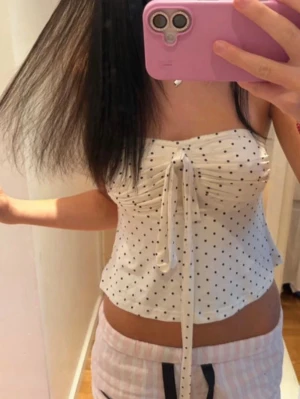 Vit prickig linnetopp med knyt - Supersöt vit linnetopp med svarta prickar och knyt framtill. Toppen är ärmlös och har en lätt croppad passform som ger en fräsch och somrig vibe. Perfekt till jeans eller kjol för en trendig look.