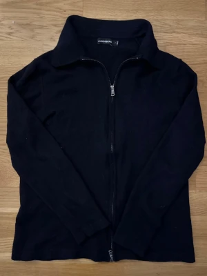 J.Lindeberg Fullzip Stickad Tröja med Krake - Klassisk svart/mörkblå ziptröja från J.Lindeberg med hög krage och hel dragkedja framtill. Tröjan har lång ärm och enkel, stilren design som passar till det mesta. Perfekt att slänga på sig när det är lite kyligare ute. Är originellt storlek L, men skulle säga mer att den sitter som S/M
