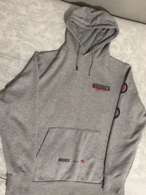 Grå hoodie från Hoodrich - Snygg grå hoodie från Hoodrich med stor logga på ryggen och mindre tryck på bröstet och fickan. Hoodien har huva med dragsko, magficka och tryck på ena ärmen. Perfekt för en streetwear-look.