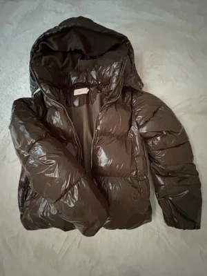 Grön pufferjacka med huva - Snygg grön pufferjacka med glansig finish och stor huva. Jackan har dragkedja framtill och två sidofickor. Perfekt för kalla vinterdagar och ger en trendig look. Materialet är syntetiskt och jackan är vadderad för extra värme.