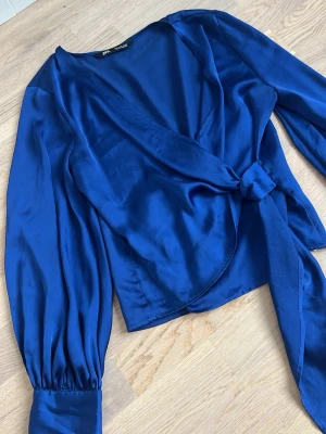 Blå omlottblus med knytband - Snygg blå omlottblus från Zara i glansigt satintyg. Blusen har långa puffärmar med breda manschetter och klädda knappar. Knytband i midjan ger en feminin siluett och djup v-ringning framtill. Perfekt för att lyfta vilken outfit som helst.