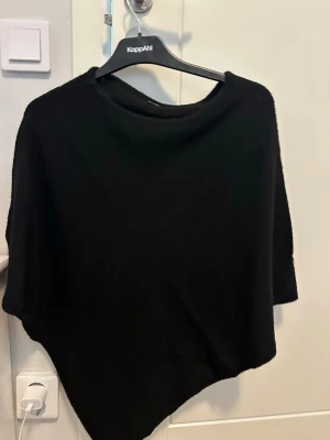 Svart stickad poncho - En svart stickad poncho med mjukt material och avslappnad passform. Ponchon har rund halsringning och asymmetrisk nederkant som ger en cool siluett. Perfekt att slänga över en tröja för extra värme och stil. Använd någon gång. Noppor förkommer 