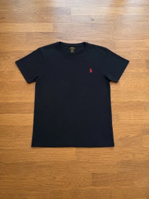 Svart t-shirt från Polo Ralph Lauren - Klassisk svart t-shirt från Polo Ralph Lauren med röd broderad logga på bröstet. T-shirten har rund halsringning och korta ärmar. Tillverkad i mjuk bomull för en skön känsla och enkel stil. Säljer billigt eftersom sommaren snart är över 