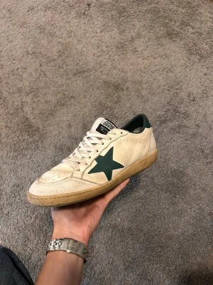 Golden Goose vita sneakers med grön stjärna - Golden Goose sneakers i vitt läder med grön stjärndetalj på sidan och grön häl. Klassisk låg modell med snörning och beige sula. Märket syns på plösen och innersulan. Perfekta för en avslappnad streetwear-look.