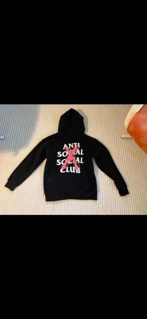 Svart hoodie med Anti Social Social Club-tryck - Svart hoodie med huva och tryck från Anti Social Social Club i vitt och rosa på både bröstet och ryggen. Klassisk passform med långa ärmar och känguruficka framtill. Perfekt för dig som gillar streetwear och vill sticka ut med en snygg logga.