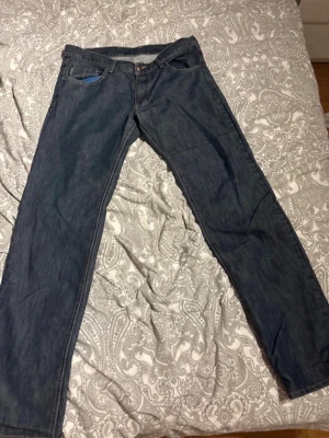Mörkblå raka jeans W36 L36 - Säljer ett par mörkblå jeans med raka ben och klassisk femficksmodell. Jeansen har kontrastsömmar och en snygg blå detalj i fickan. Perfekta för dig som gillar enkel och stilren denim.