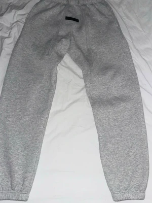 Grå mjukisbyxor från Essentials - Snygga grå mjukisbyxor från Essentials med resår i midjan och benslut. Byxorna har dragsko i midjan och en svart logotyp framtill. Tillverkade i mjukt och bekvämt material, perfekta för en avslappnad stil.